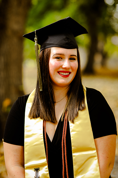 Victoria is posing for a picture wearing a graduation cap and her sash and ropes to indicate her top academic performance. She is smiling and looking directly at the camera.