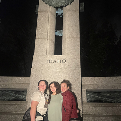 Victoria, Kellie and Maddie stand together, posing, in front of a tall granite column that says Idaho. Click to enlarge.