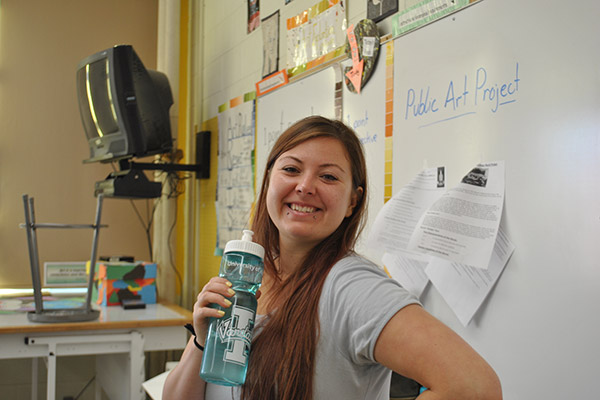 Kalli smiles and poses for the camera while standing at the front of a classroom. She is holding a water bottle.