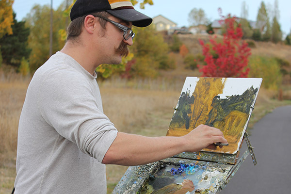 Aaron Johnson is standing at an easel outside, thoughtfully painting the landscape around him with oil paints on a canvas