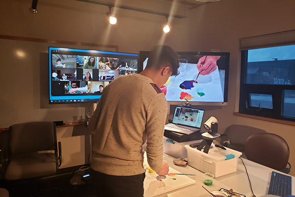 Ernesto is standing in front of two large screens and a camera. He is demonstrating a painting technique while participants look on over Zoom.