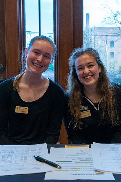 Julia is sitting on the left, next to another young woman. Julia is dressed in all black, smiling and looking at the camera.