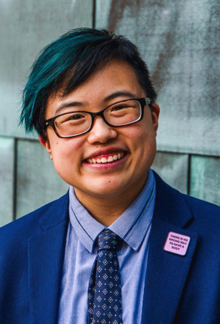 Lydia is standing in front of a brick wall and smiling happily. They have short black hair, died blue at the front. They are wearing black-framed glasses, a blue blazer, light blue button-down shirt and a navy-blue tie.