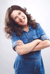 Rosie is standing playfully with her arms crossed. She is smiling and wearing a denim jumpsuit. Her long brown hair is lose around her shoulders.