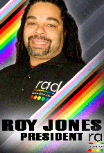Roy is smiling directly at the camera. He is a black man, with long, black dreadlocks pulled away from his face. He also has a moustache and goatee and he is wearing a black polo shirt. His image has a rainbow effect running diagonally in the background and foreground.