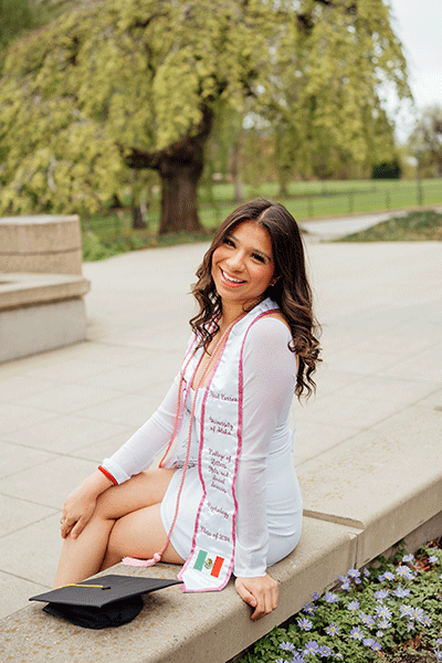 Abril is sitting on a ledge, looking over her shoulder at the camera. She is wearing a white dress with sheer sleeves and a white sash to celebrate her Academic Certificate. She is smiling and her long dark hair is loose around her shoulders.