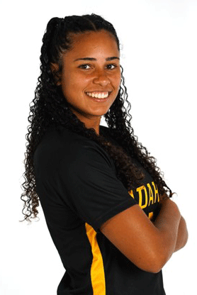 Maya is standing in front of a white background. She has her arms crossed and is looking over her shoulder at the camera. She is smiling, with her dark, curly hair pulled back away from her face in a half up-do. She is wearing a University of Idaho soccer jersey.