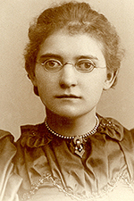 Agatha is looking directly at the camera with a neutral facial expression. Her hair is tied up into a bun and she is wearing a dark dress with a high collar.