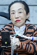 Alice is looking directly at the camera. She is sitting in her wheelchair with her BiPap machine. She is smiling slightly. She has short, black hair slicked back and is wearing red lipstick along with a tiger-striped sweater.