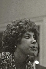 This is a profile picture of Joyce. She is a person of color, learning forward and looking intently at whatever is in front of her. Her face is serious. She has short, curled hair and is wearing a patterned blouse.