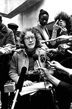 Kitty is sitting in a room in her wheelchair. There are reporters all around her, holding microphones to her face. She is wearing glasses and looking slightly down at the microphones as she speaks.