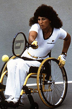 Marilyn is playing tennis in her Quickie wheelchair. She is intently looking at an oncoming ball as she swings her racket toward it. She has long, permed, brown hair and is wearing white althletic clothing.