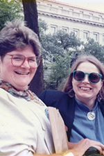 Mary Lou and Patrisha are sitting together on a bench outside. Both are smiling happily as they look at the camera. Mary Lou (left) is wearing large red-framed reading glasses and a white jacket. She has short, brown hair. Patrisha is wearing big, round sunglasses. She is wearing a blue jacket and has shoulder-length, brown hair.