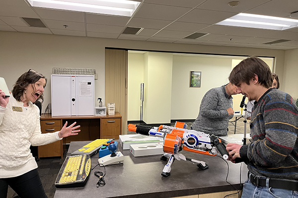 Caleb is standing behind a Nerf toy and pretending to push the button that will shoot a Styrofoam bullet while a woman stands in front of him and feigns surprise. 