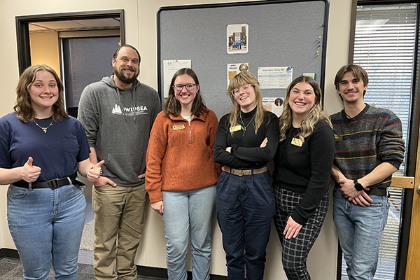 Five student trainees, four young women and a young man, are standing in a group with a male SWITC staff. All are smiling and looking at the camera. The young woman on the left end is giving two thumbs up.