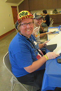 Mike is sitting at a table during an artAbility workshop. He is wearing a blue t-shirt, baseball hat and glasses. He is looking directly at the camera and smiling. Mike is holding a blank canvas up, with an art tool in his hand, preparing to get started on his project.