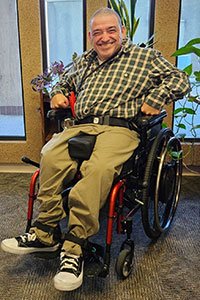 Noll is sitting in a wheelchair in a brightly-lit room. He is smiling broadly for the camera. Noll is wearing a tan flannel button up shirt and khaki dress pants. He has very short, dark hair.