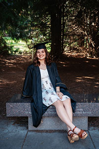 Isabella is sitting on a bench on the UI campus with trees around her. She is wearing a black graduation gown over a white knee-length dress with a blue floral print. She is also wearing a graduation cap and is smiling with her dark blonde hair falling around her shoulders.