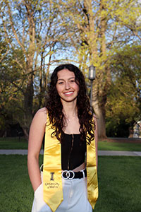 Jayda is standing on a green lawn on the UI campus with trees in the background. She is smiling and has her hands in the pockets of her white slacks. She is wearing a black tank top with a gold University of Idaho sash around her shoulders. Her black, curly hair is falling around her shoulders.