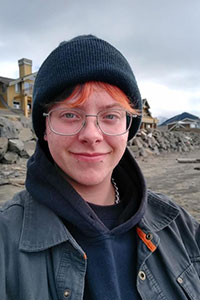 Kincaid is standing on a beach on an overcast day. He is smiling at the camera with rocks in the background. He is wearing a black beanie, glasses and a dark hoodie under a dark blue jacket.