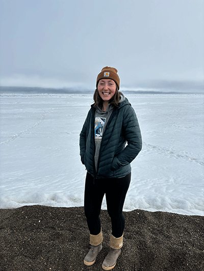 Grace is standing on a snowy field. There are heavy clouds behind her. She is smiling broadly, wearing a beanie, heavy coat, black pants and boots.