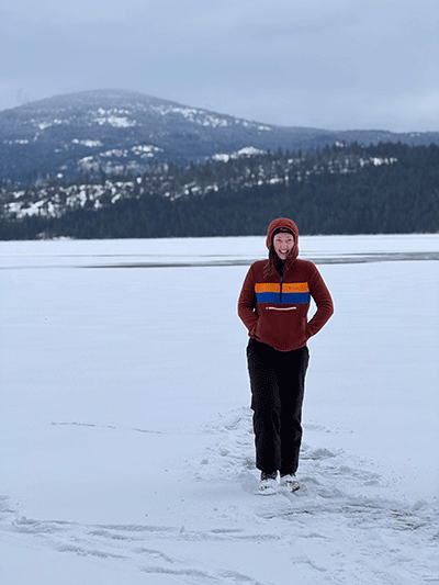 Grace is standing in the middle of a cold, snowy landscape with a mountain behind her. She is bundled up in a heavy coat with a hood on and is smiling happily.