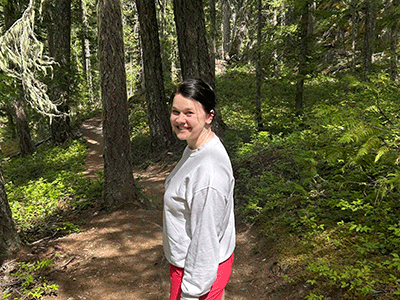 Suzanna is walking along a trail through the forest. There are evergreens all around her. She is looking back and smiling happily at the camera. Her brown hair is pulled into a pony tail and she is wearing a grey sweatshirt and bright pink shorts.