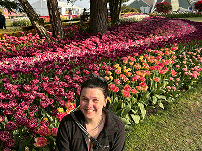 Suzanna is sitting in front of a large bed of tulips. They are all different colors and in full bloom behind her. She is smiling happily. Her dark hair is pulled back into a ponytail and she is wearing a black jacket.