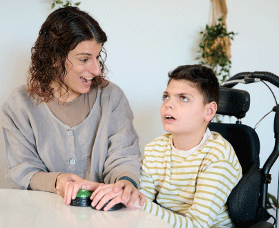 A woman sits with a young boy with a disability. She is helping him use adaptive assistive technology switches.