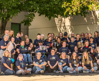 A group of young people with disabilities gathered with graduate students and support staff for a group photo. Everyone is wearing the same AAC camp shirt. They are all smiling and looking at the camera.