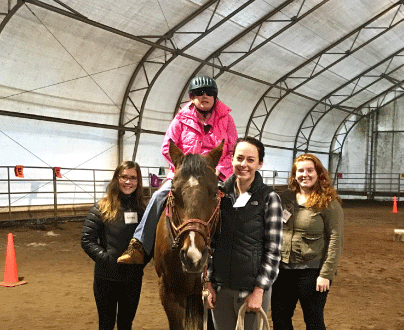 A young woman holding a horse by its reigns. She is smiling at the camera. A young girl with vision loss is on the horse, and two other women are standing on either side of the horse. Both are smiling at the camera.
