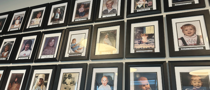 A wall of framed photographs. Each photo is of a young child or baby. They each have the name of the person pictured below the image. Each is smiling at the camera or playing in their photo.