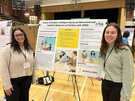 Two young women are standing on either side of their presentation poster about their research project. They are in a large gymnasium with other presentation posters from other students. They are smiling proudly.