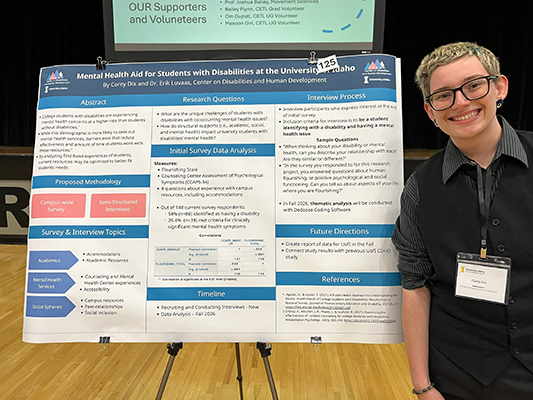 A student trainee is standing next to their presentation poster about their research project. They are in a large gymnasium with other presentation posters from other students. They are smiling proudly.