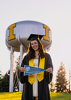 Avery is standing in front of the iconic University of Idaho water tower. She is wearing her graduation garb and holding a pennant for the Florida grad school she will be attending.