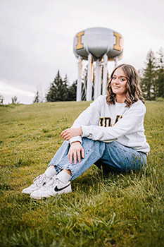 Haley is sitting on the lawn in front of the iconic University of Idaho water tower. She is wearing a University sweat shirt and smiling directly at the camera.