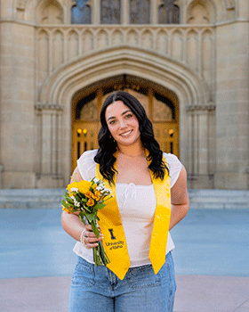 Natalie is standing in front of the University of Idaho Admin building. She is wearing a gold university sash around her neck and holding a bouquet of flowers. She is smiling happily at the camera.