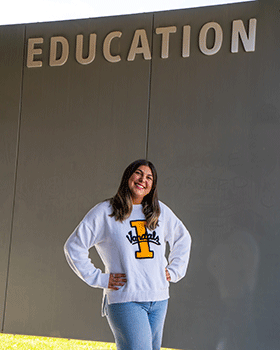Sofia is standing under a sign on a building that says Education. She is smiling and has her hands on her hips. She is wearing a University of Idaho sweater.
