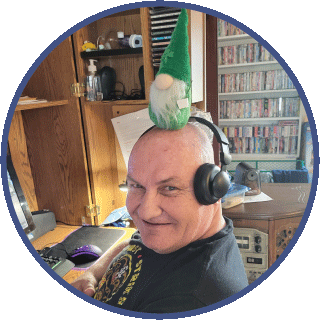 Sam is looking directly at the camera. He is balancing a miniture green gnome on his head and smiling mischeviously.