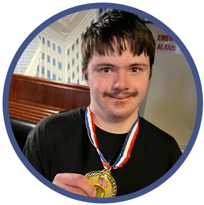 Gabe is smiling slightly while looking at the camera. He is standing and holding a gold medal that is hanging around his neck.