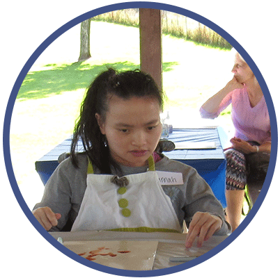 Hannah is sitting at a table outside, working on a piece of art. She is looking at her art, with her hands on either side of her canvas. She is wearing an apron over a grey, long-sleeved shirt. She has black hair that it tied back into a ponytail.