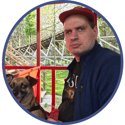 Peter is looking at the camera, wearing a red cap and blue jacket. He is sitting with his dog in an outdoor area.