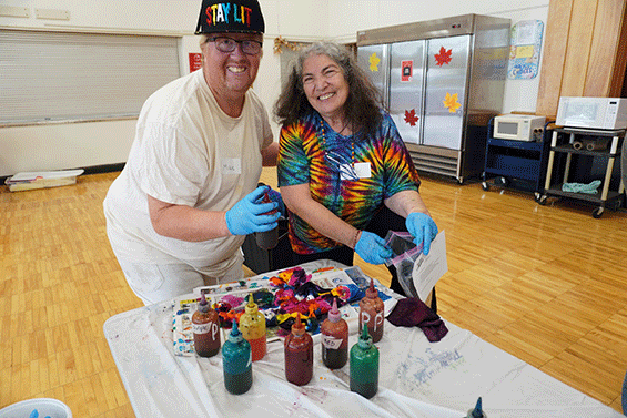A male self-advocate is standing next to an older woman who is showing him how to tie dye an apron. They are both looking at the camera and smiling happily.