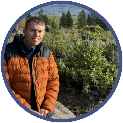 Carter is standing outside, with wilderness behind him. He is looking at the camera and smiling. He is wearing a puffy orange and blue winter coat.