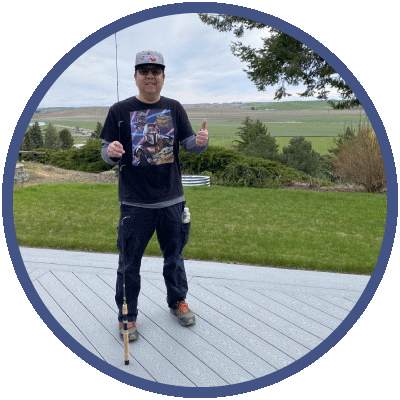 Cory is standing outside. He is wearing a hat and sunglasses, and holding a fishing pole. He is smiling slightly and giving a thumbs up to the camera.