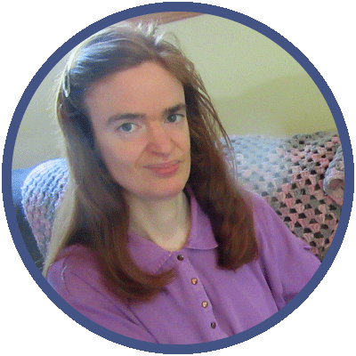 Deirdre is sitting on a chair. She is looking at the camera and smiling slightly. She has long red hair and is wearing a purple polo shirt.