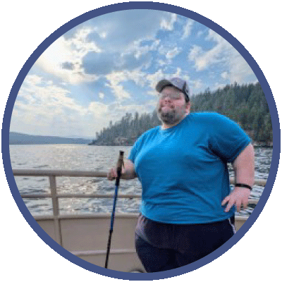 Erik is standing on a boat in the water. There is an expanse of water behind him, framed by mountains covered in evergreens. Erik has one hand on his hip and is smiling slightly at the camera. He is wearing a hat, glasses and blue t-shirt.