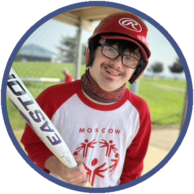 Gabe is standing outside at a baseball diamond. He is wearing a batting helmet and holding a baseball bat. He is smiling happily. He has glasses and dark brown hair.