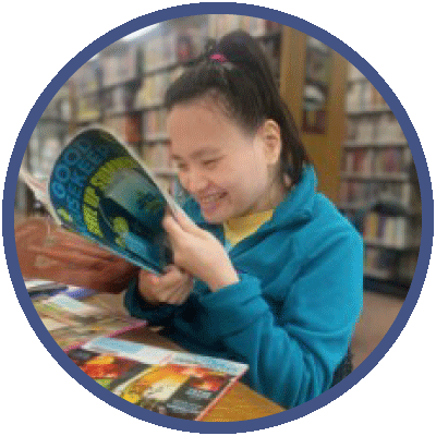 Hannah is sitting at a table in a library. She is reading a magazine with a big smile on her face. She has short, dark hair that is pulled up away from her face and she is waring a yellow t-shirt under a blue jacket.