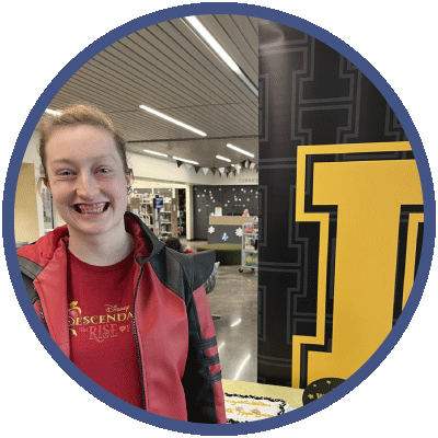 Hope is standing next to a large yellow University of Idaho I. She is smiling broadly. Her hair is pulled back away from her face and she is wearing a red t-shirt under a red and black jacket.